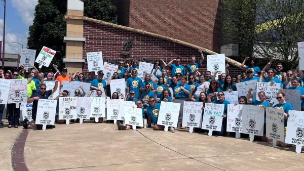 A large group of ASF Mankato members in teal shirts and Teamsters Local 320 representatives standing shoulder-to-shoulder outside a campus brick building, holding rally signs advocating for fair pay and workplace equity.